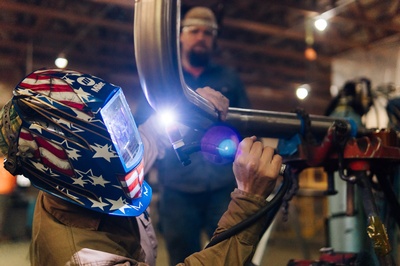 welder in stars and stripes helmet works with a torch on a curved metal pipe while being observed.