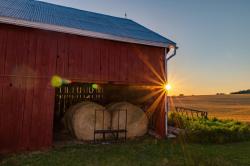 Sunlight peeks out along the edge of a red barn with two large hay bales showing in a wide opening