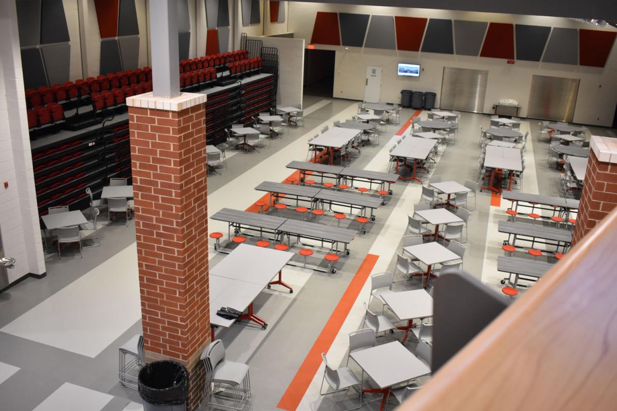 Red and black bleachers are retracted against the wall and cafeteria seating is lined along the open eating space. 