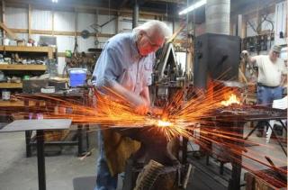 An older man is working at a modern blacksmith forge. Sparks fly out.