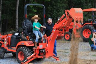 a boy in a cowboy hat sits operates the claw on a large Kubota tractor with an adult supervising