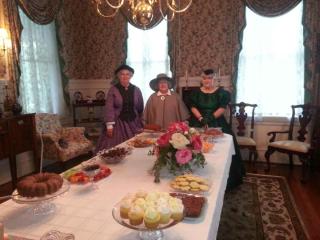 3 ladies in period dress stand behind a table loaded with goodies