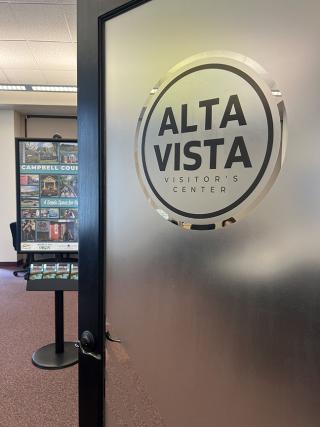 A frosted glass door with "Altavista Visitor Center" inscribed opens into a room with a poster stand showing County photos