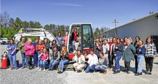A group of around 30 women gather around a mini-excavator and give a "thumbs up." Two women sit in the drivers seat area.