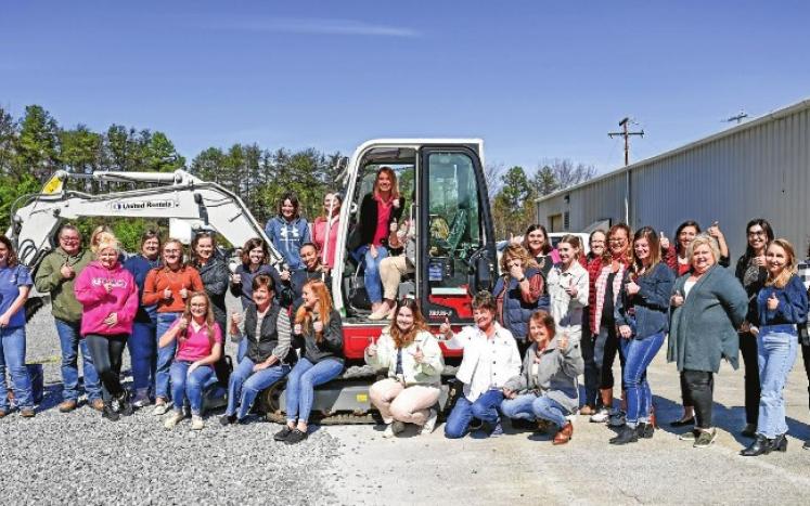 A group of around 30 women gather around a mini-excavator and give a "thumbs up." Two women sit in the drivers seat area.